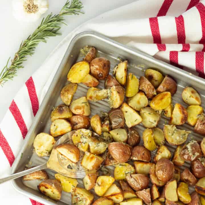 A sheet pan of oven roasted baby potatoes on a red and white striped napkin. A sprig of rosemary and a head of garlic are in the background.