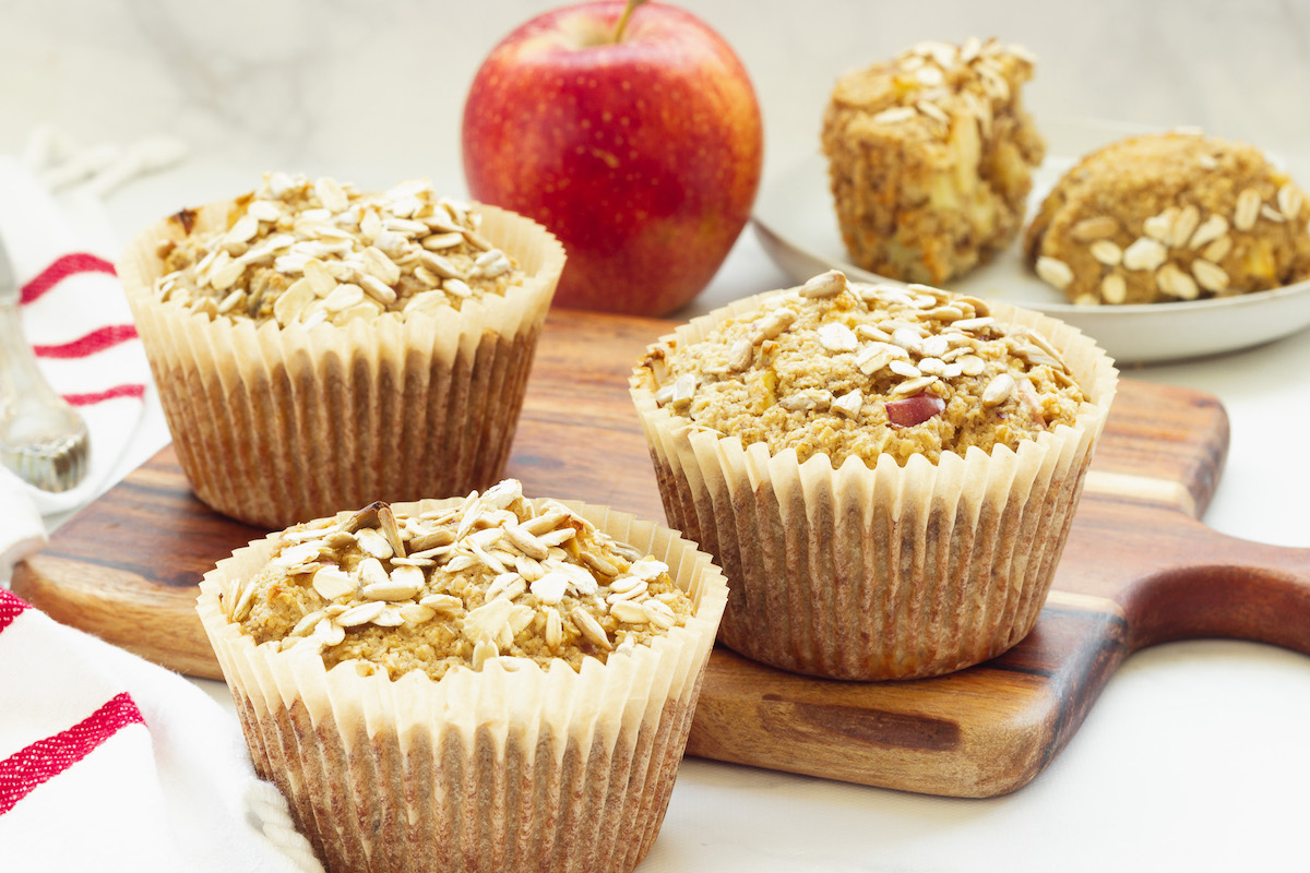 Four oat bran muffins and an apple sitting on a cutting board.