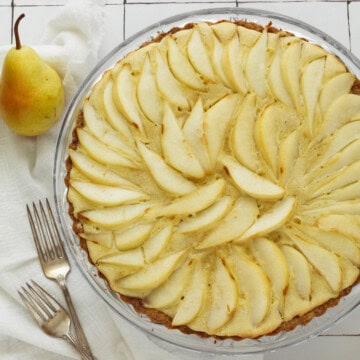 A baked pear tart with two forks, a white napkin, and a pear next to the tart pan.