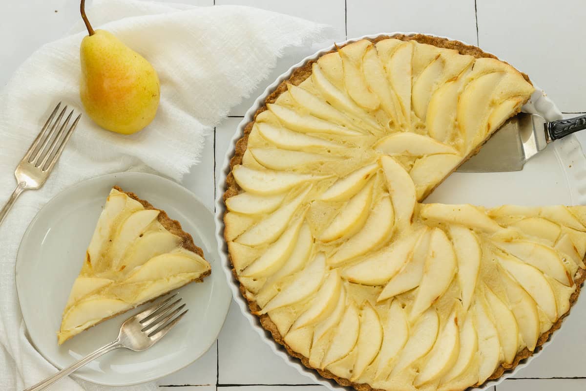 A pear tart with one slice one slice on a white serving plate. A pear and white napkin are beside the tart.