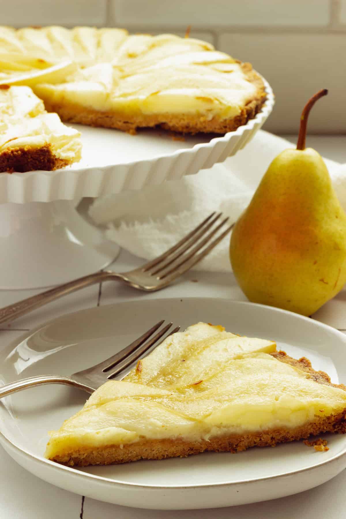 A slice of baked pear tart on a white plate with a fork. A tall dessert plate with the pear tart is in the background.