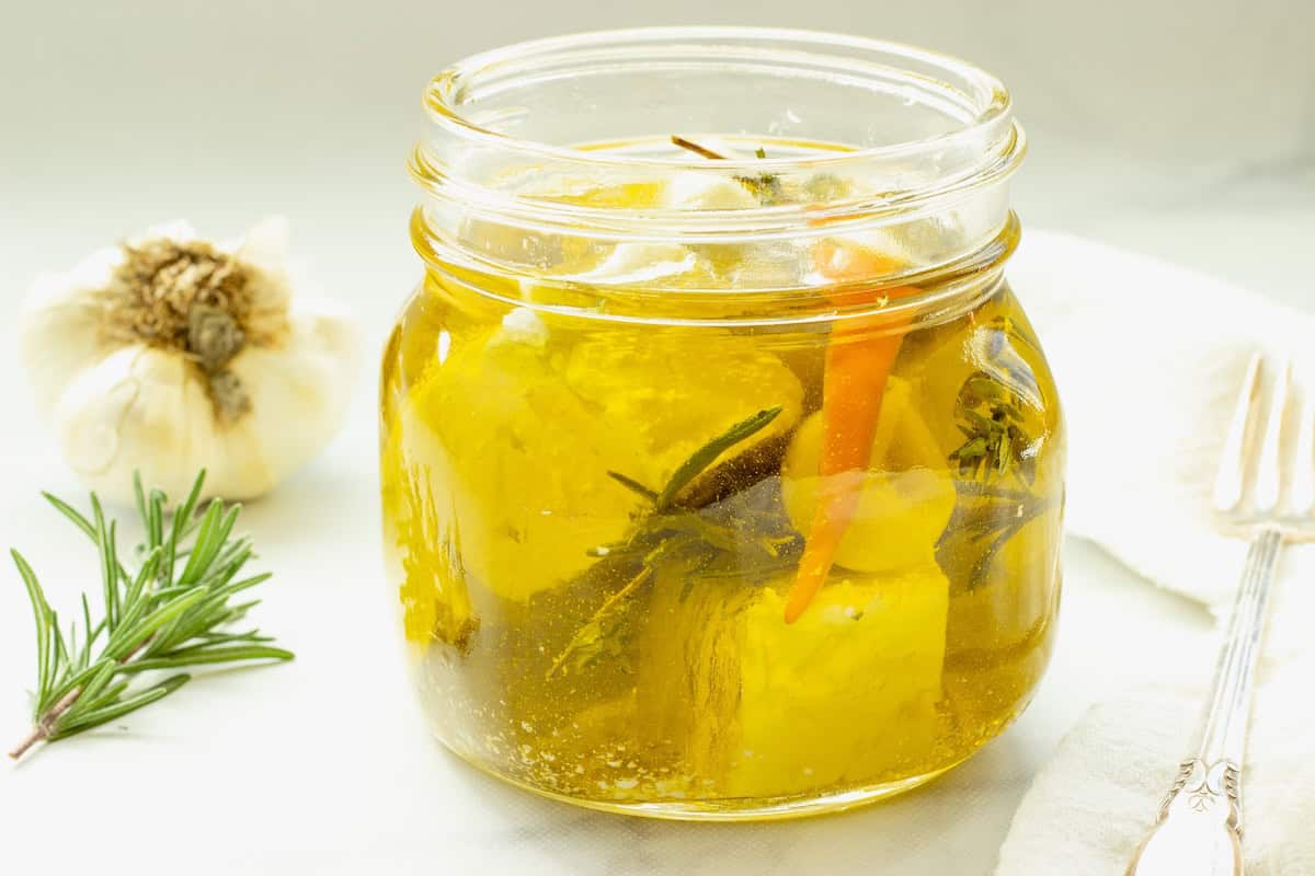 A jar of marinated feta cubes. A head of garlic and spring of rosemary are next to the jar.
