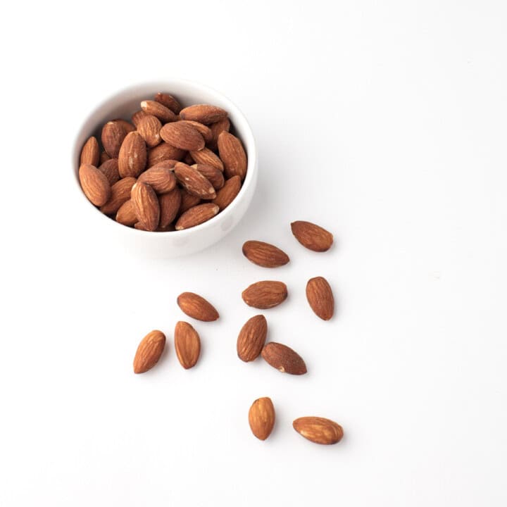 a white bowl of almonds with a handful of loose almonds lying next to the bowl.