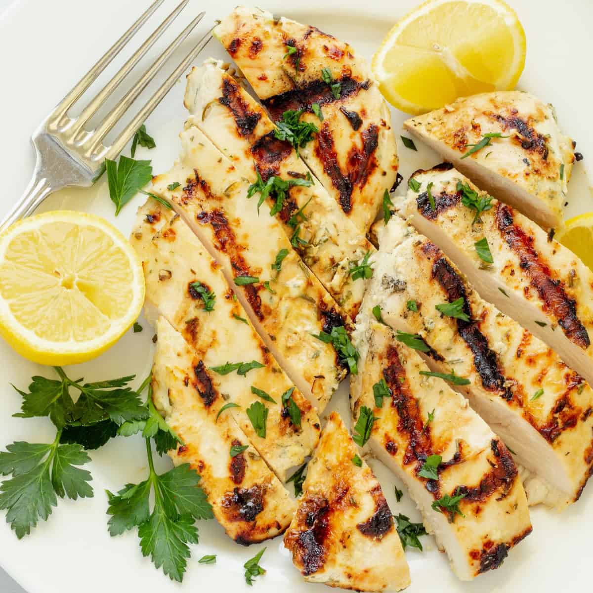 A platter of sliced Greek yogurt marinated chicken with lemon slices and parsley.