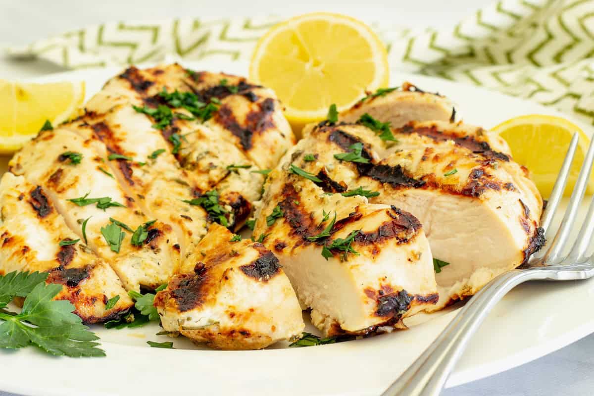 A white platter of sliced grilled yogurt marinated chicken with lemons, parsley, and a fork alongside the chicken.