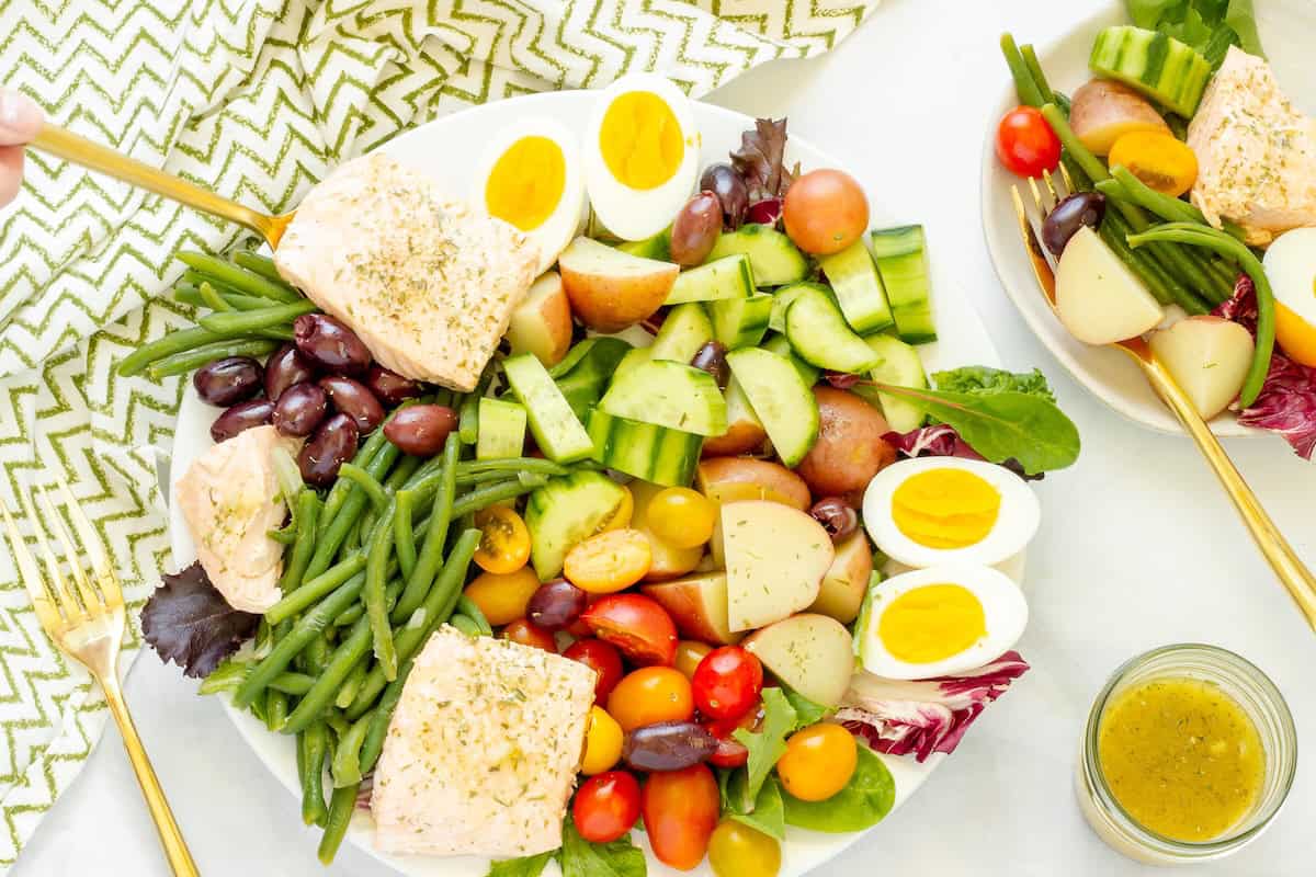 A composed platter of salmon Nicoise salad and a serving of the salad on a white table. Gold serving utensils and a green and white patterned napkin are on the table.