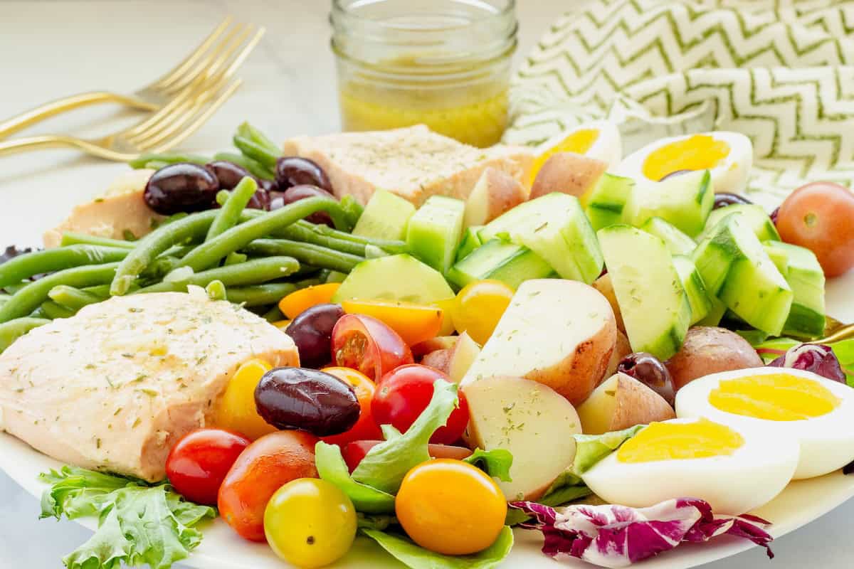 A composed platter of salmon Nicoise salad. A bottle of salad dressing and gold serving spoons are alongside the plate.