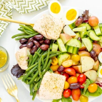 A composed platter of salmon Nicoise salad with a green and white pattern napkin and serving utensils.
