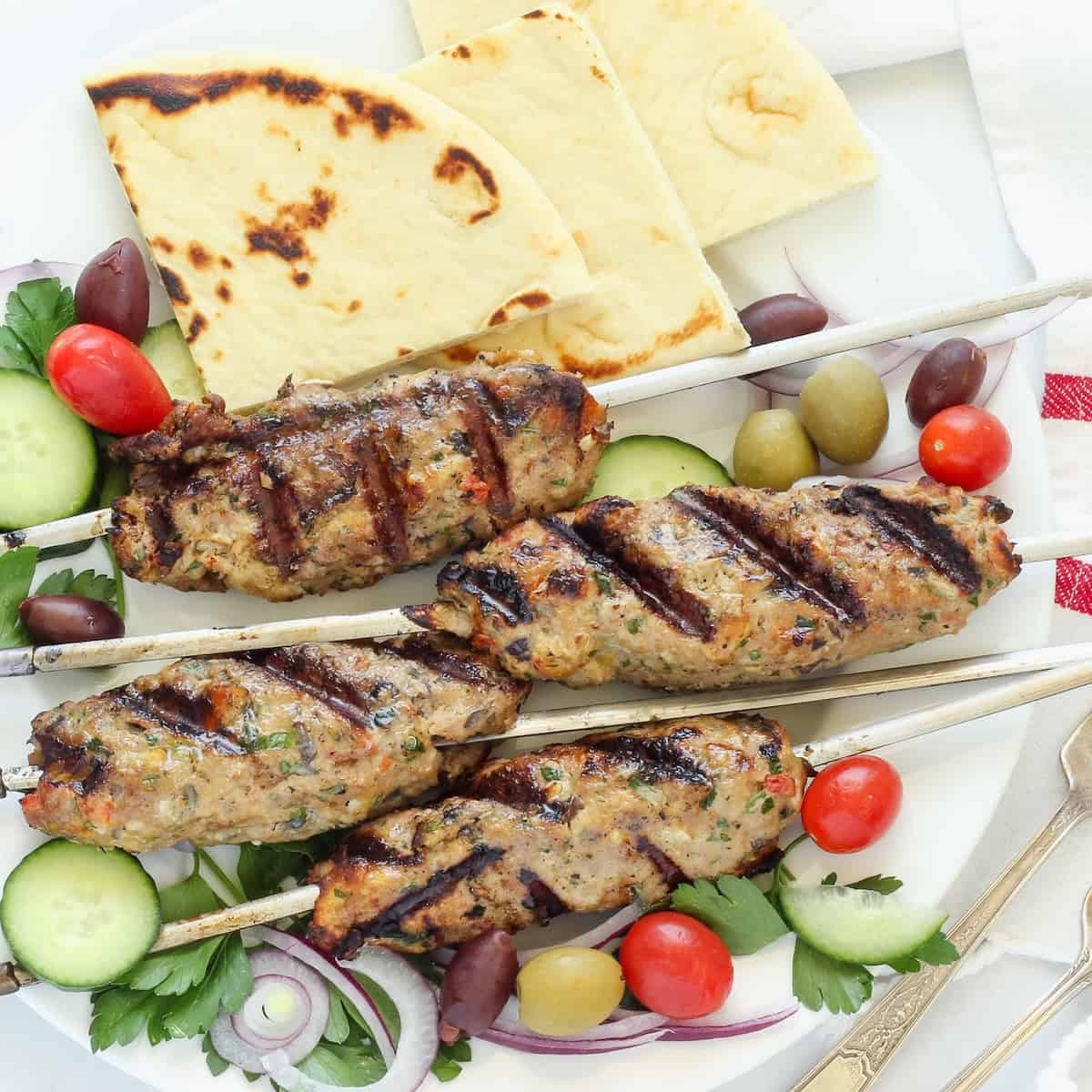 A platter of chicken kofta garnished with cucumber tomato salad, olives, and pita bread.