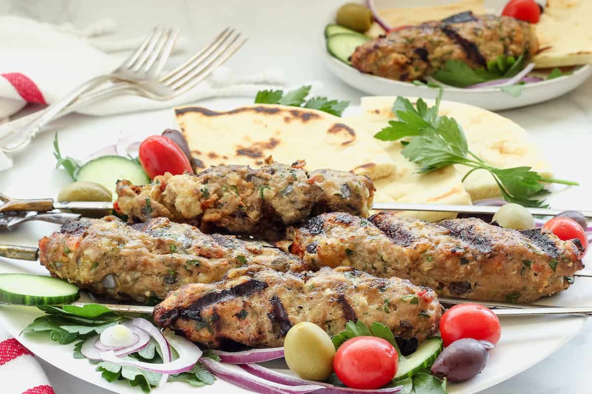 Chicken kofta skewers garnished with olives, tomatoes, parsley, and pita bread.