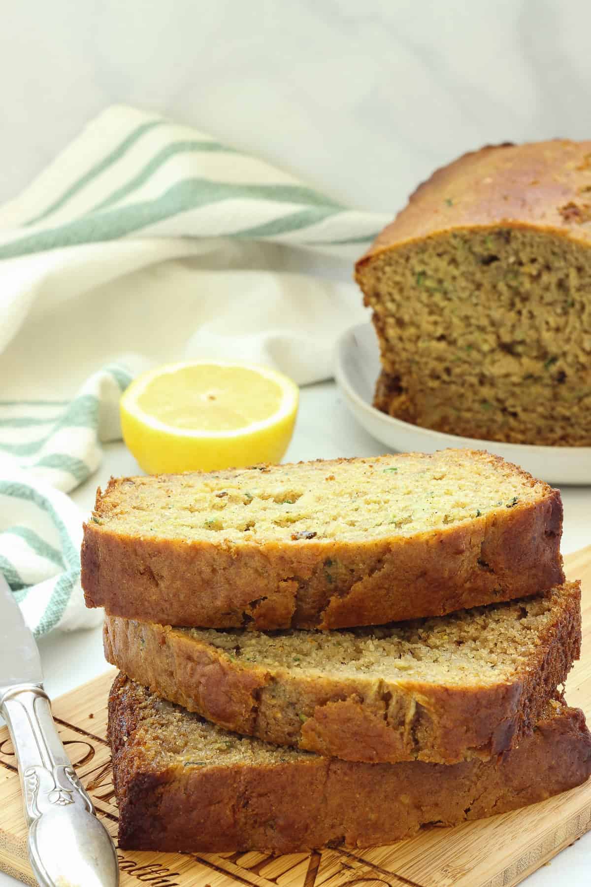 Stacked slices of healthy lemon zucchini bread next to a green and white striped napkin, a cut lemon, and half a loaf of the bread.